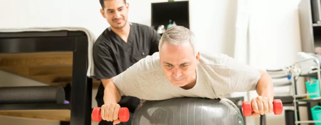 Patient Lifting Dumbbells While Lying On Exercise Ball By Physiotherapist
